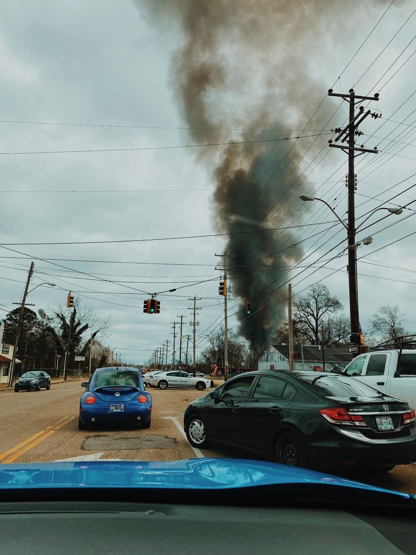 This is a photo taken from the inside of a blue car. The photo is of a plume of smoke in the distance and there is a black sedan and a blue volkswagen bug being directed by the police away from the smoke. In the distance there is a Memphis Police Car in the distance under the streetlight. The sky is a light blueish, grey and the street is brown.