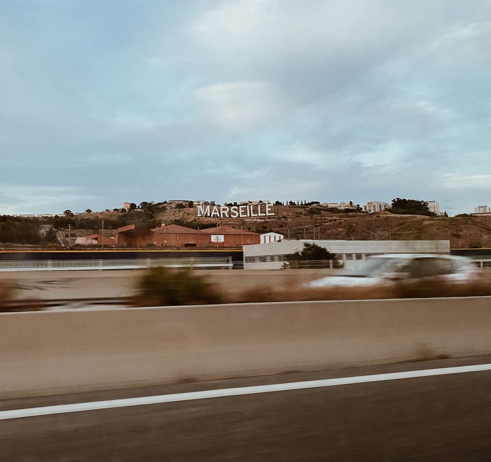A photograph of a French landscape, taken from the window of a moving vehicle. The huge white letters of MARSEILLE are in the center of the piece. Blurred vehicles pass underneath the signage, and a light blue sky is clustered with gray clouds.