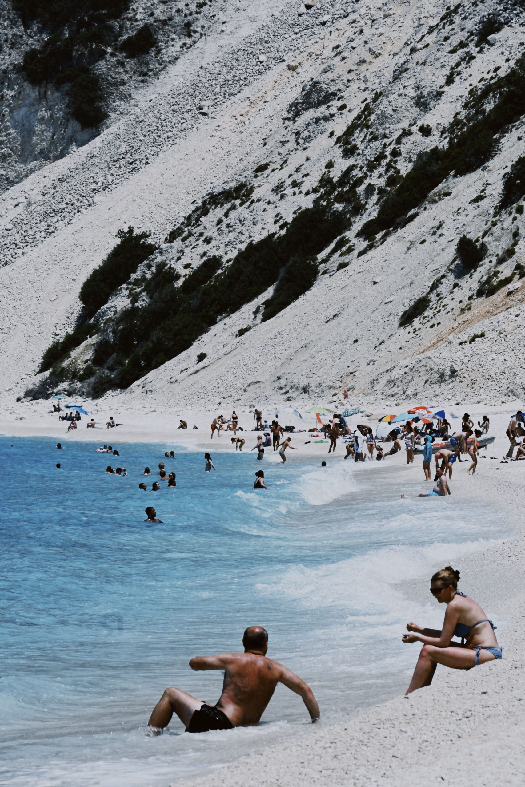 Teal blue water cuts into the white sands of a beach. The background of the image is sloping sand that cuts diagonally into the distant water. The sand slope goes up so high that you cannot see the sky at all. There are people playing in the distant shore, and a couple sits in the water close to the camera, the woman facing the waves and the man in the water, reaching back to her.