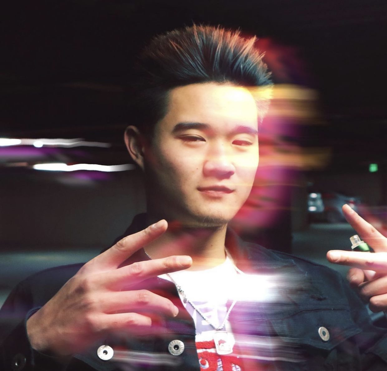 This is a photograph of a man with peace signs and a vape in his hand. The light and color on his face is smeared and the colors are saturated. The smearing makes the image appear to be in motion although the man in the photo is stationary. The man is wearing a black jacket and white shirt. He has black pointy hair as well.
