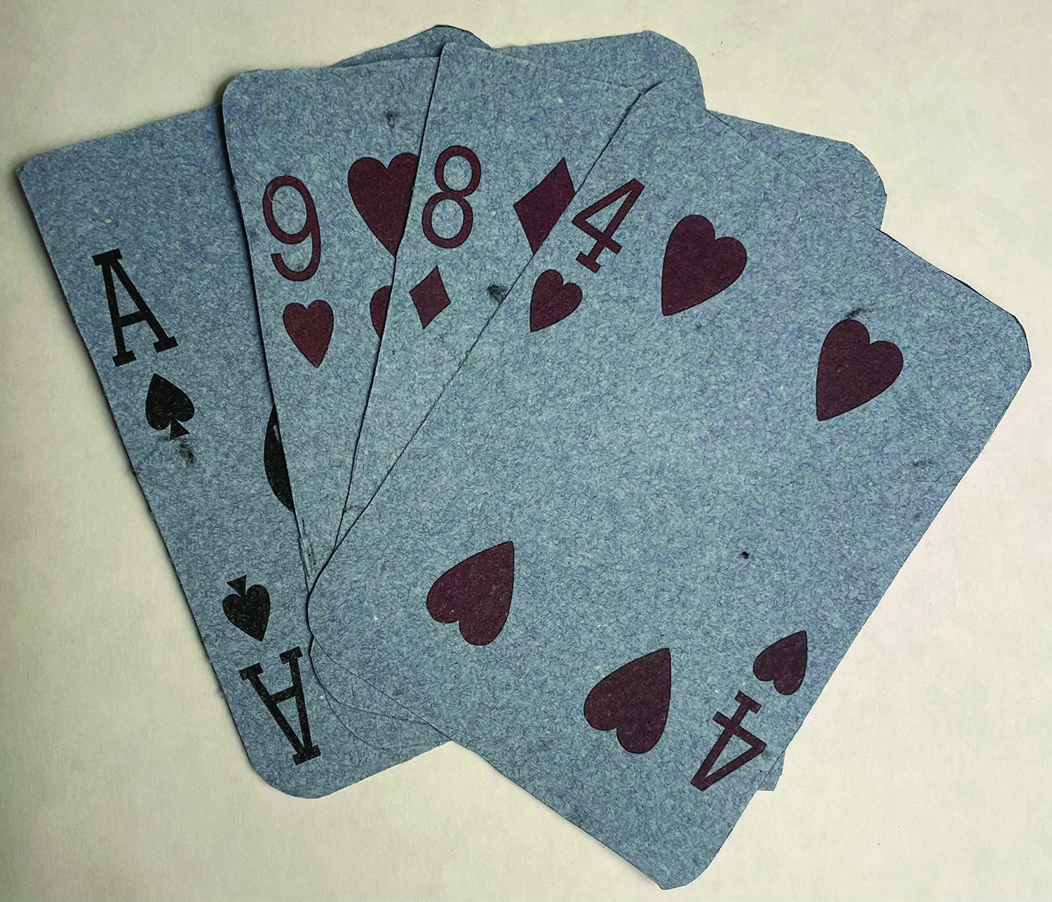 Four playing cards made from handmade paper consisting of pulp made from blue button-down shirts. The cards are fanned out, with a red four of hearts in the forefront. It is followed by a red eight of diamonds, a red nine of hearts, and a black ace of spades as the final card. The pulp texture from the handmade paper is present on the surface of each card. They are placed against a beige background.