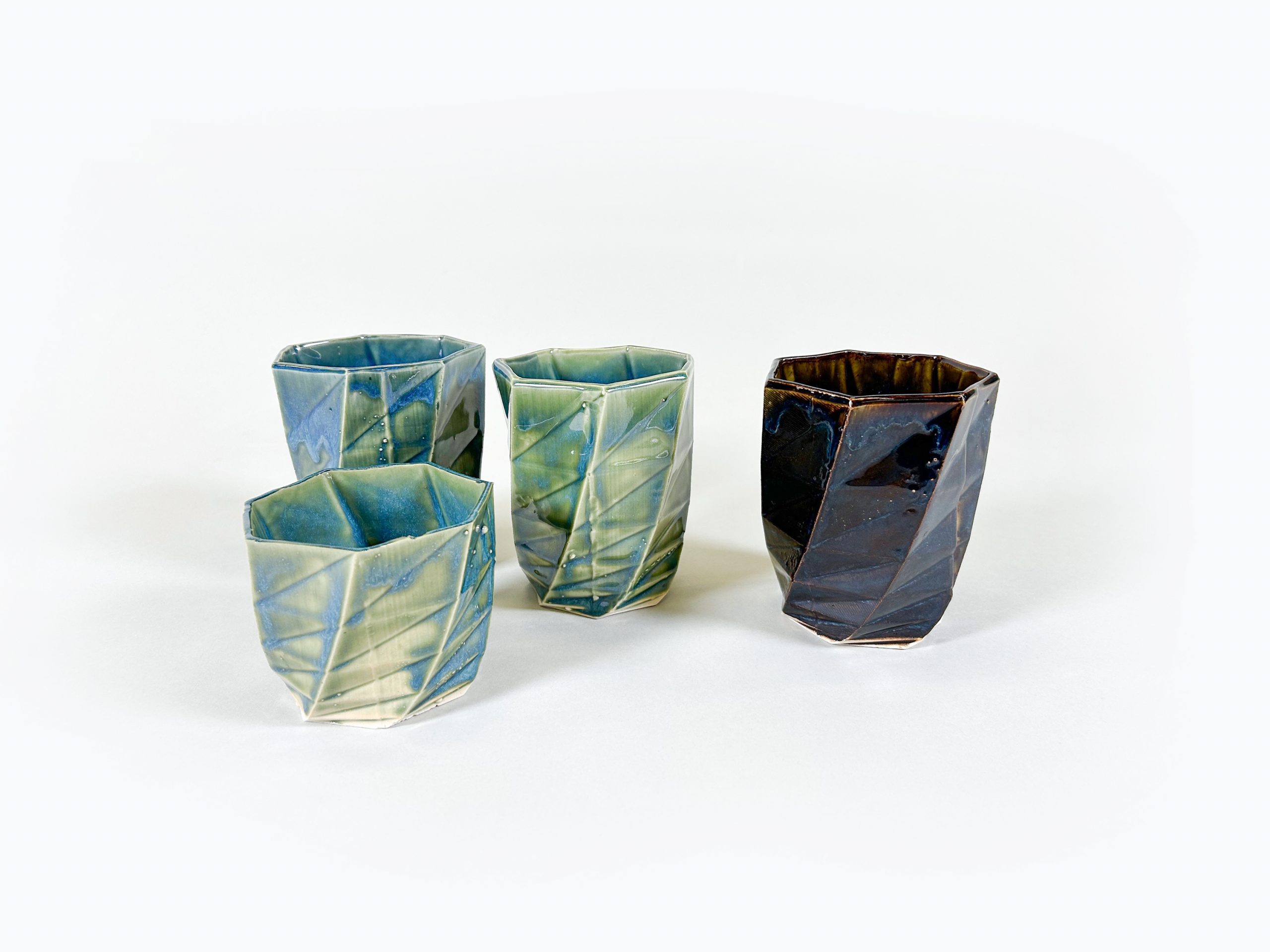 A collection of four porcelain cups created using slip-casing and 3D modeling. One smaller cup sits in the foreground while three taller cups of the same size sit behind it. The rims of the cups are seven-sided polygons which appear to rotate the length of one side as the walls approach the base of the form. Three cups, including two tall cups and the one short cup, are glazed in a translucent green color, with blue crystals forming along the lines of the cups and within certain thicker areas of glaze. The final large cup is a deep cholate brown. On this cup, dark blue crystals creep out from the lines within the cup, turning to white at the edges.