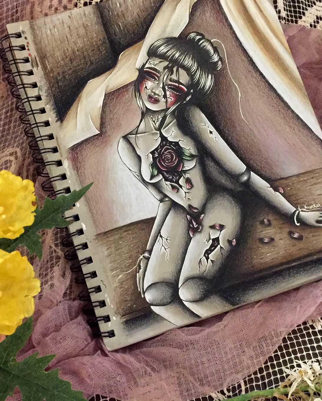 A illustration in a sketchbook of a broken marionette puppet, of mostly grays and blacks, sitting on a brown table. A large gaping wound in the doll's chest opens to reveal a rose of dark red petals, deep green leaves, and white highlights. Petals fall from the rose into the lap of the puppet. The doll's head is tilted toward the right, limp, and her black hair is tied in a loose bun with strands falling in front of her face, slightly covering the upside-down crescent moon symbol on her forehead. There are broken strings hanging from the wrists and head of the marionette. The sketchbook is pictured on a background of light pink and white lace fabrics, with a pop of bright yellow flowers with green leaves on the left side.