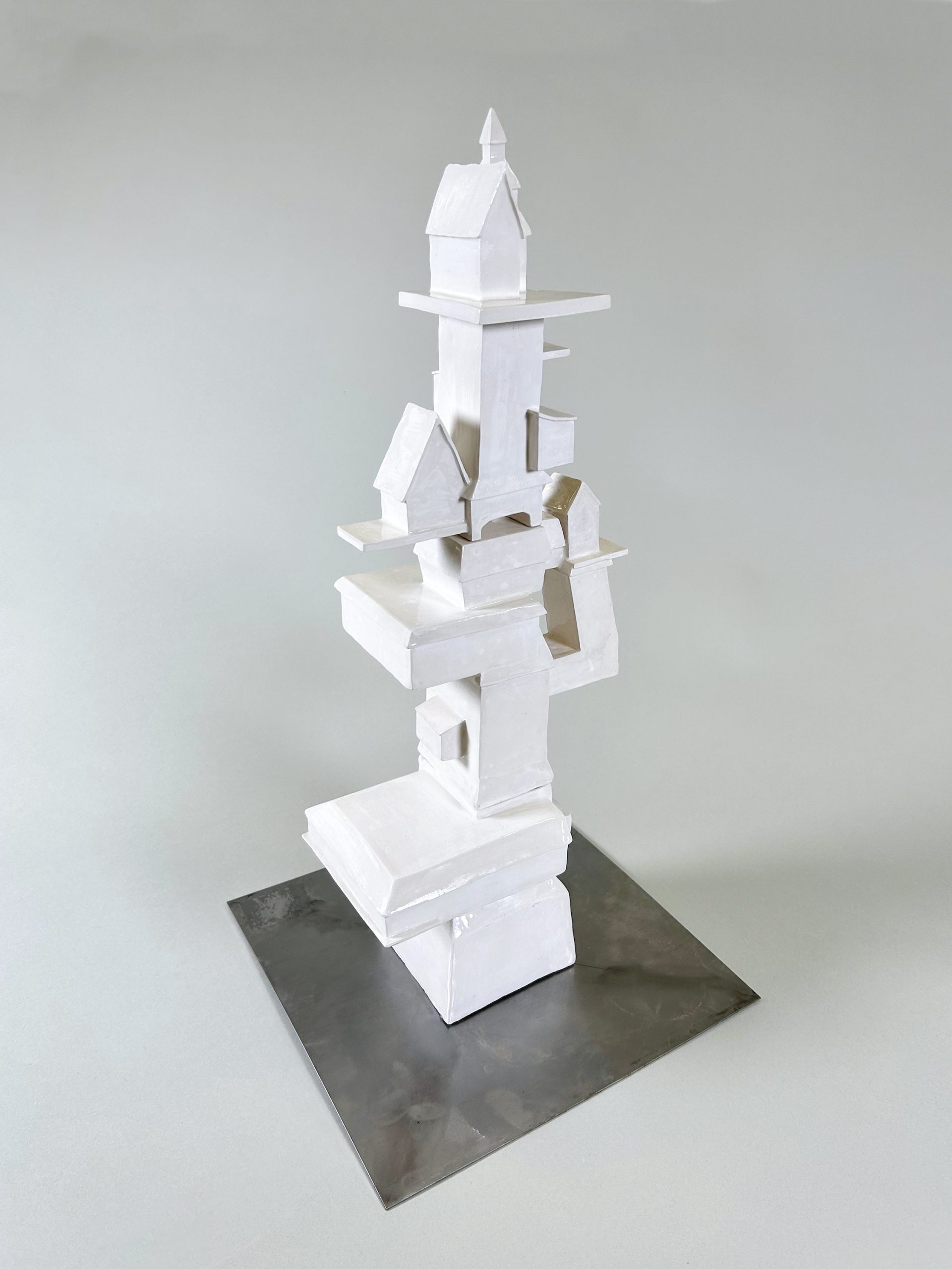 A 2-foot tall white, ceramic tower standing at a 3-quarters angle. It has a 1-foot-wide block as a foundation and becomes narrower as it approaches the top. It is topped with a house-like structure with a triangular roof. The tower has three structures that come out on the sides, attached diagonally on the opposite side of the tower. The bottom-most addition is a flat box. the middle-most is a perpendicular arm that upholds a small house-like building. Not far above is a medium-sized house-like structure on a thin base. the artist says. "